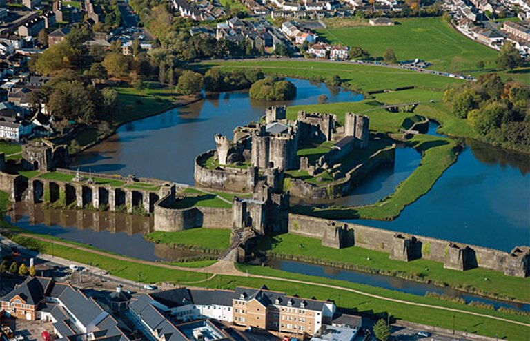 An aerial view of Caerphilly Castle, a massive medieval stone fortress surrounded by concentric rings of water and extensive green lawns. The castle complex, with its numerous towers and walls, is centrally located within the image. Surrounding the castle are modern residential areas with houses and green fields. The sky is clear with soft light.