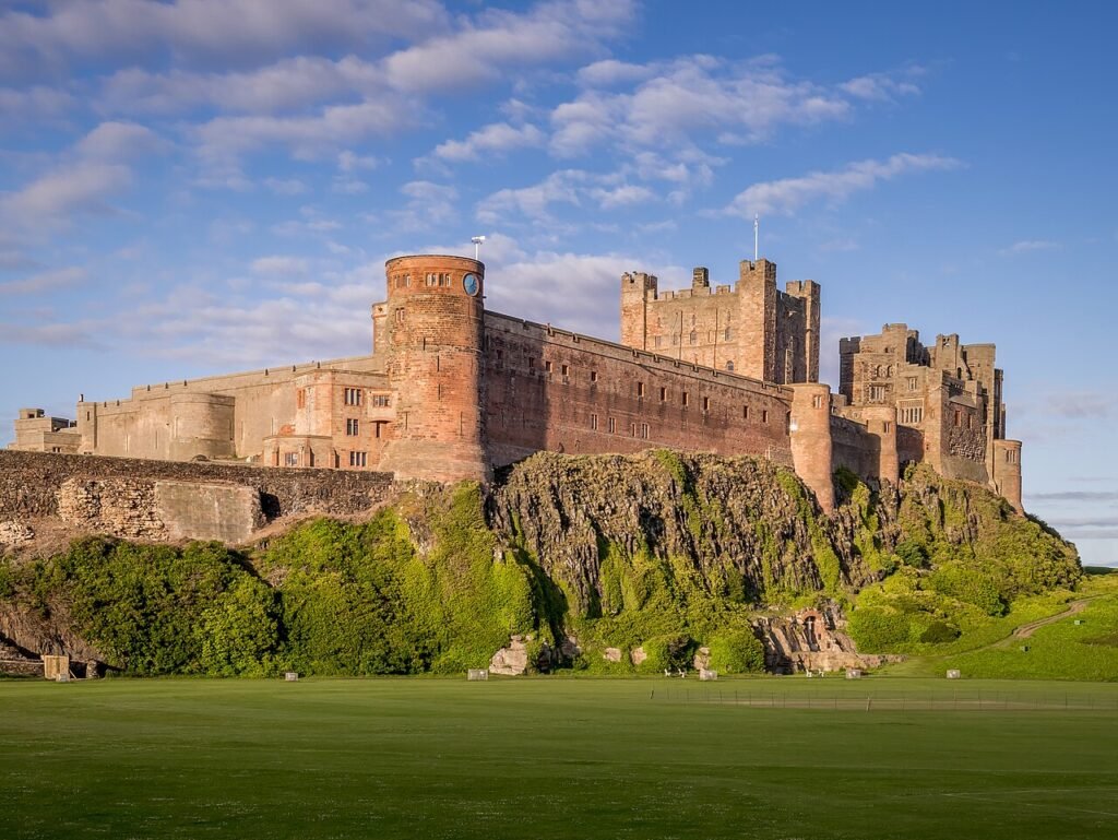 Bamburgh Castle, a large stone fortress with reddish-brown walls, standing on a rocky cliff above a green lawn under a blue sky with white clouds.
