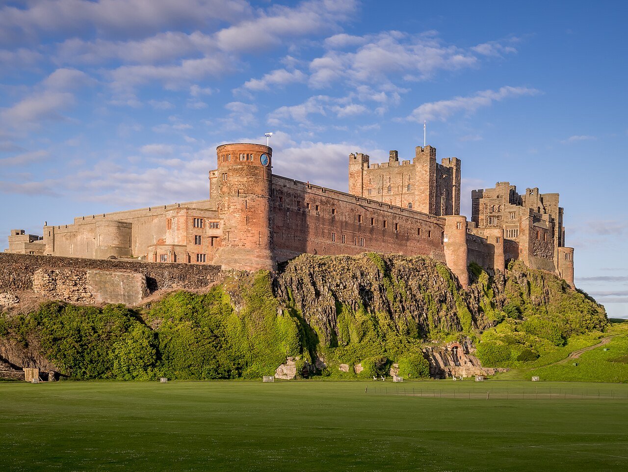 Bamburgh Castle, a large stone fortress with reddish-brown walls, standing on a rocky cliff above a green lawn under a blue sky with white clouds.