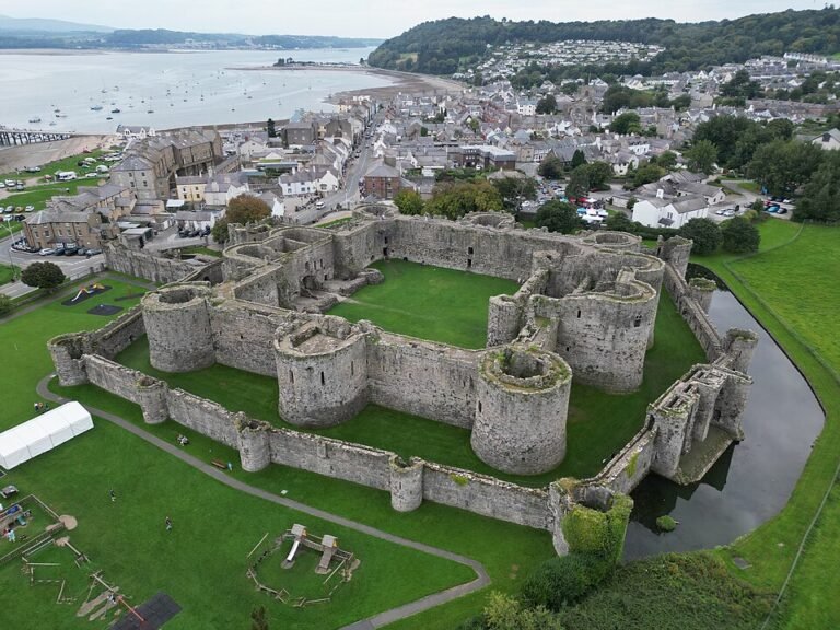 An aerial view of Beaumaris Castle, a large concentric medieval fortress with a central green courtyard, surrounded by two rings of grey stone walls and a moat, on the island of Anglesey, Wales. The castle is surrounded by manicured green grass. Beyond it, the town of Beaumaris with its grey and white buildings stretches towards a wide bay dotted with boats. Hills and a cloudy sky form the distant background.