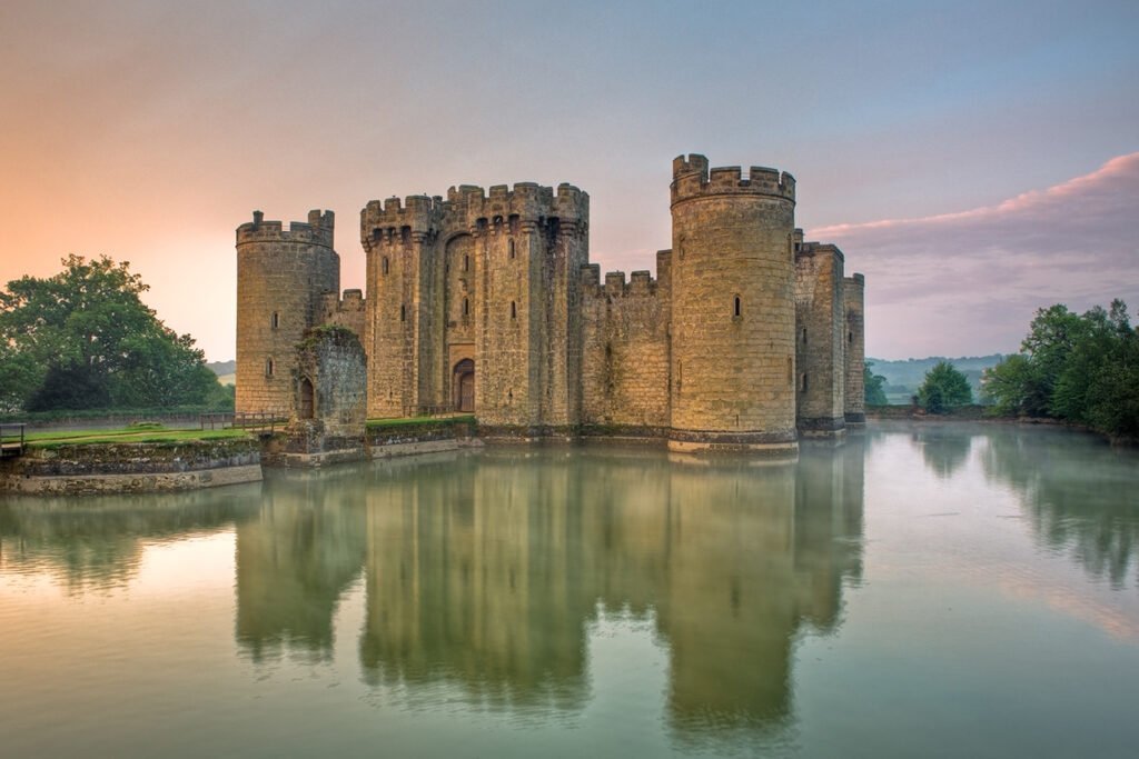 A view of Bodiam Castle, a medieval stone fortress with round towers, surrounded by a wide, reflective moat under a pastel sky.