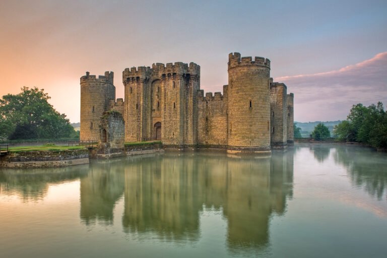 A view of Bodiam Castle, a medieval stone fortress with round towers, surrounded by a wide, reflective moat under a pastel sky.