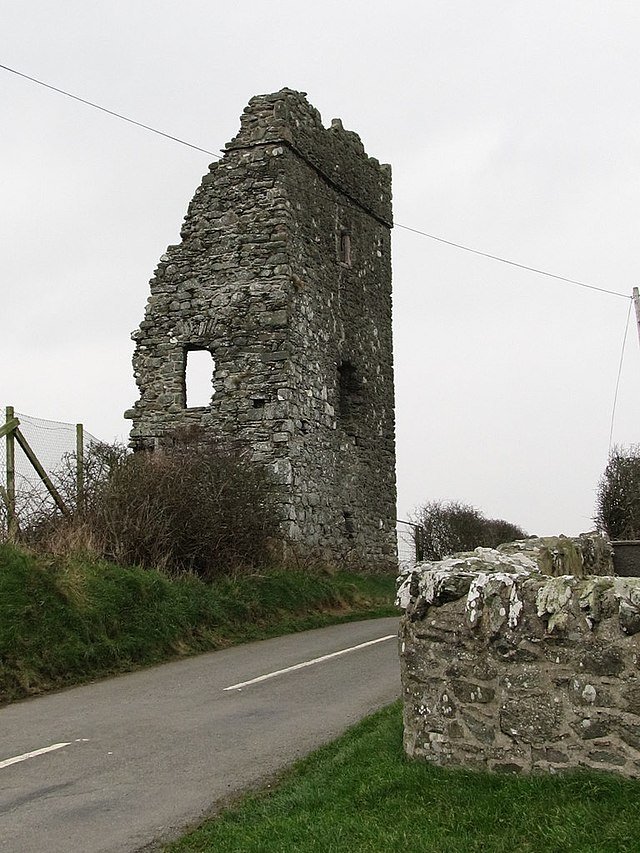 Bright Castle: A photo of the stone ruins of Bright Castle, showing a tall, angular section of a grey stone tower, with several window openings and a jagged top, standing next to a narrow paved road. A low stone wall is in the foreground, and a wire fence and overgrown bushes are behind the ruin. The sky is overcast and grey.