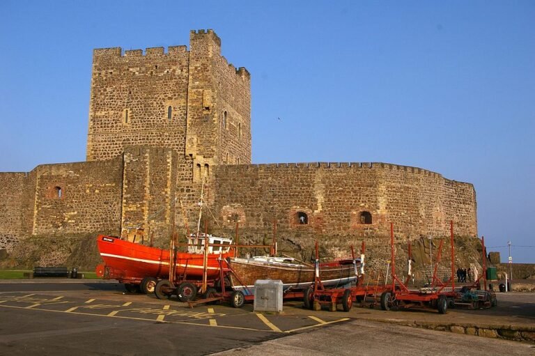 Carrickfergus Castle: A large, medieval stone castle with high walls and a square keep, located on a rocky coast with several fishing boats on trailers in the foreground.