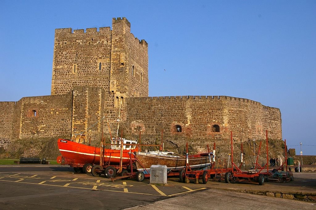 Carrickfergus Castle: A large, medieval stone castle with high walls and a square keep, located on a rocky coast with several fishing boats on trailers in the foreground.