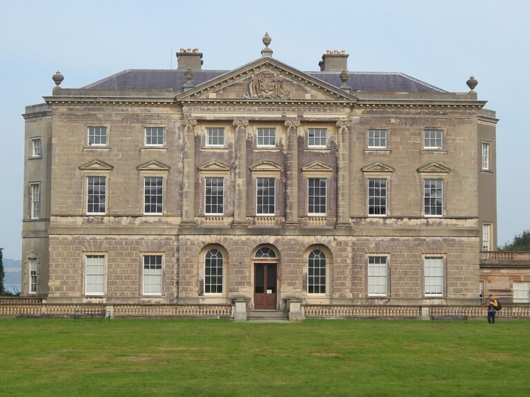Castle Ward: A frontal, symmetrical shot of Castle Ward, a large country house with a grey stone facade. The building has a central, pedimented section with large columns and multiple large windows on two floors. The lower level has a set of arched windows and a central doorway. The house is surrounded by a well-maintained green lawn. A small person is visible on the right side of the frame, providing scale.