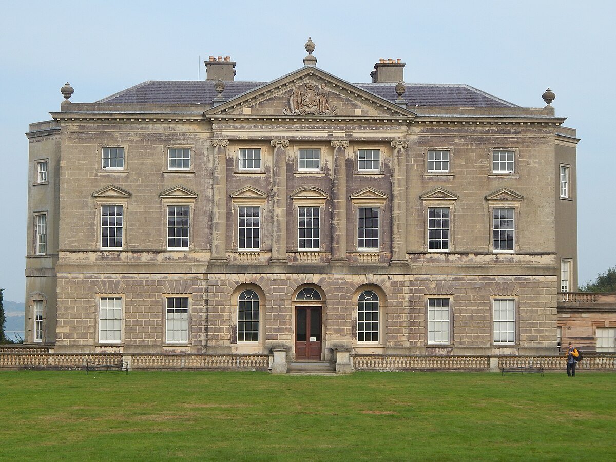 Castle Ward: A frontal, symmetrical shot of Castle Ward, a large country house with a grey stone facade. The building has a central, pedimented section with large columns and multiple large windows on two floors. The lower level has a set of arched windows and a central doorway. The house is surrounded by a well-maintained green lawn. A small person is visible on the right side of the frame, providing scale.
