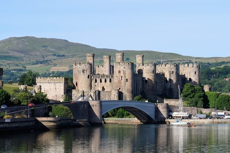 A scenic view of Conwy Castle, a large medieval stone fortress with multiple tall, round towers and extensive walls, situated on the edge of a body of water. A white arched bridge spans the water in front of the castle. Hills with green and brown foliage rise in the background under a clear blue sky. Several boats are docked on the right side of the water.