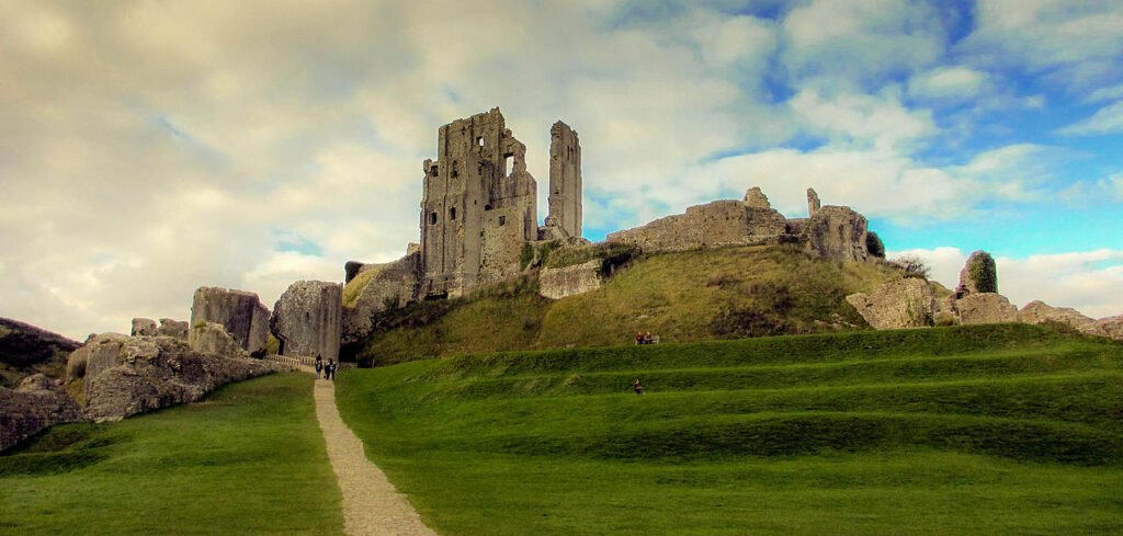 Corfe Castle: The dramatic ruins of a medieval stone fortress on a grassy hill, with a path leading up to the broken towers and walls under a cloudy sky.