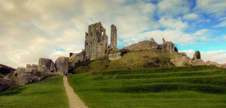Corfe Castle: The dramatic ruins of a medieval stone fortress on a grassy hill, with a path leading up to the broken towers and walls under a cloudy sky.
