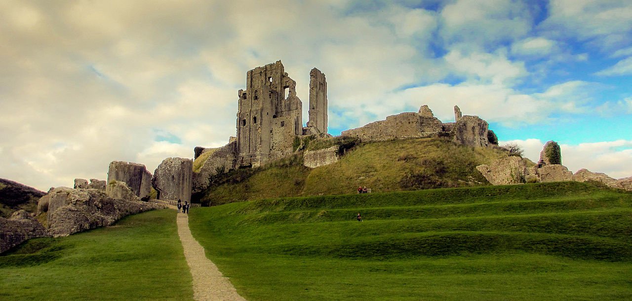 Corfe Castle: The dramatic ruins of a medieval stone fortress on a grassy hill, with a path leading up to the broken towers and walls under a cloudy sky.