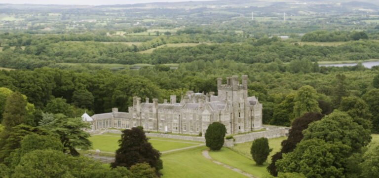 An aerial view of Crom Castle, a large, grey stone Victorian castle with many turrets and battlements, nestled amidst dense green woodland. A manicured lawn and pathways surround the castle. In the distance, a vast expanse of green countryside with a body of water is visible under a light grey sky.