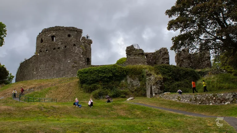 Dundrum Castle: A photo of the stone ruins of Dundrum Castle on a grassy hill under a cloudy sky. The main visible structure is a large, round keep with some windows and battlements. Other fragmented walls and overgrown sections are also visible. Several people are scattered around the grassy pathways and slopes, some walking and some sitting, exploring the historic site. A large tree with dark leaves is on the right.