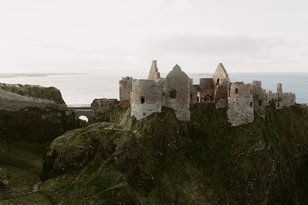 A photo of the ruins of Dunluce Castle perched on a rocky cliff overlooking the sea. The stone walls of the castle are mostly intact, but the roofs and some of the upper floors are missing. A small stone bridge connects the castle to the mainland. The sky is overcast and the sea is calm.