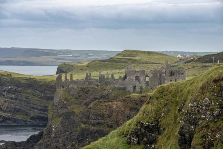 Dunluce Castle: A distant, horizontal shot of the ruins of Dunluce Castle on a grassy headland. The castle is a long, low structure with jagged walls and missing roofs. It sits on a cliff overlooking the sea. The surrounding landscape is hilly and green, with a few white buildings visible on the horizon. The sky is cloudy and grey.