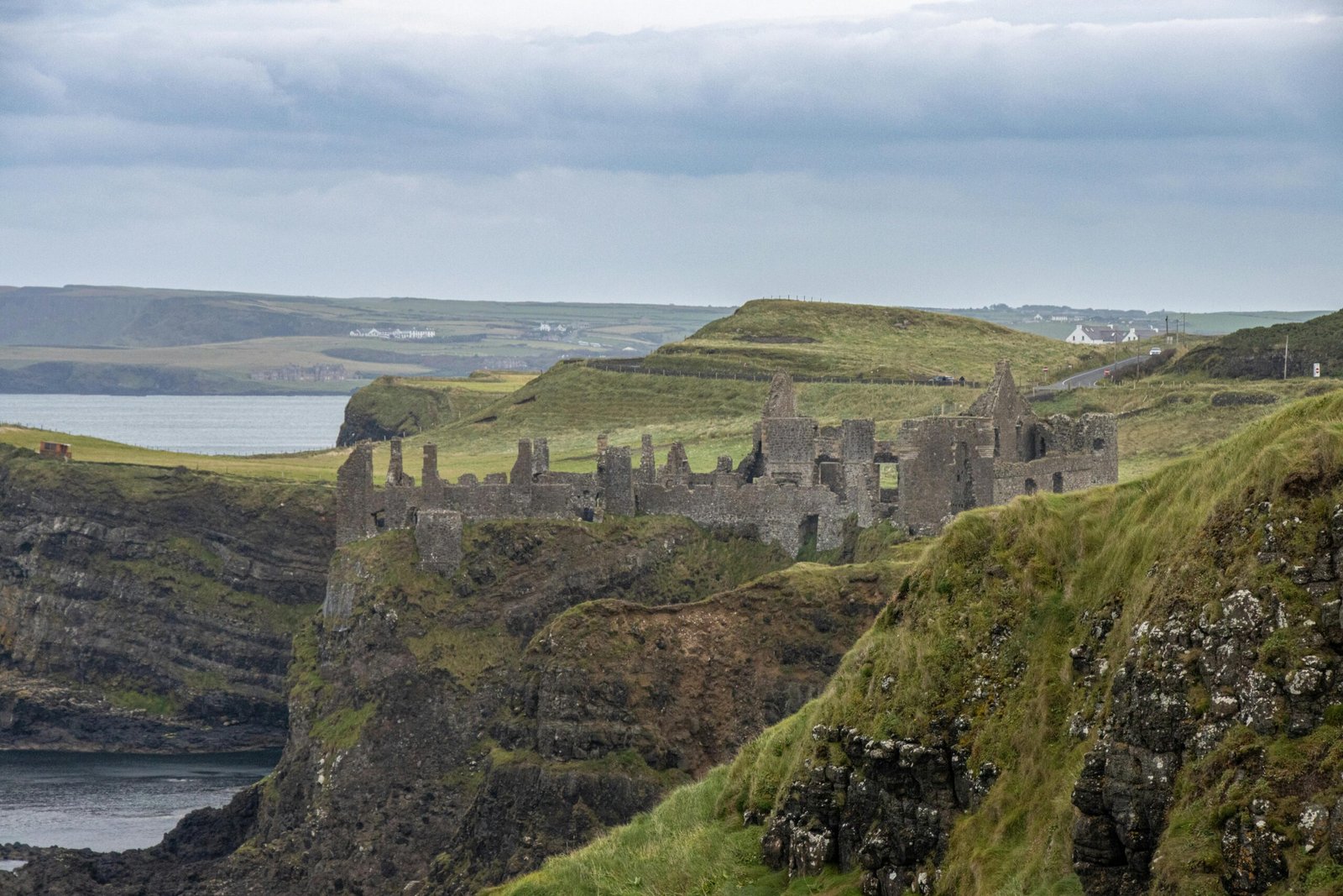 Dunluce Castle: A distant, horizontal shot of the ruins of Dunluce Castle on a grassy headland. The castle is a long, low structure with jagged walls and missing roofs. It sits on a cliff overlooking the sea. The surrounding landscape is hilly and green, with a few white buildings visible on the horizon. The sky is cloudy and grey.