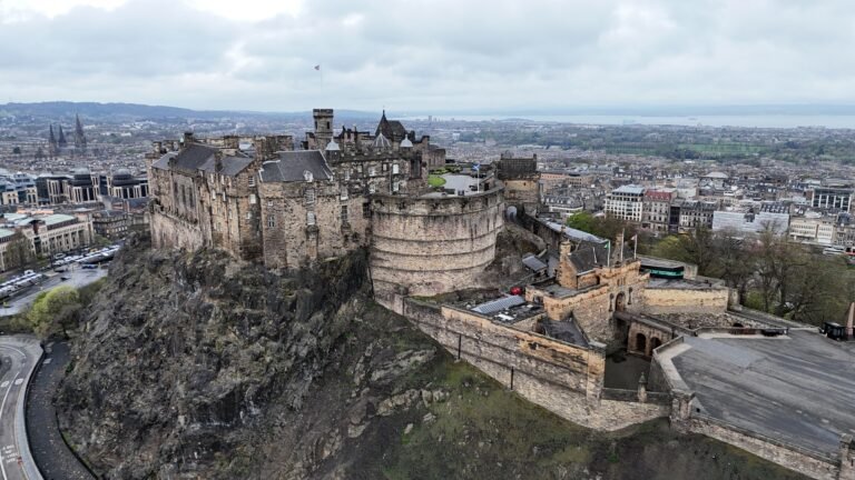 An elevated view of Edinburgh Castle, a large, historic stone fortress with multiple buildings, towers, and walls, perched on a rugged volcanic rock. The castle dominates the left side of the frame. Below and around it, the city of Edinburgh stretches out, with numerous grey buildings and streets. A body of water, likely the Firth of Forth, is visible in the distance. The sky is overcast and grey.