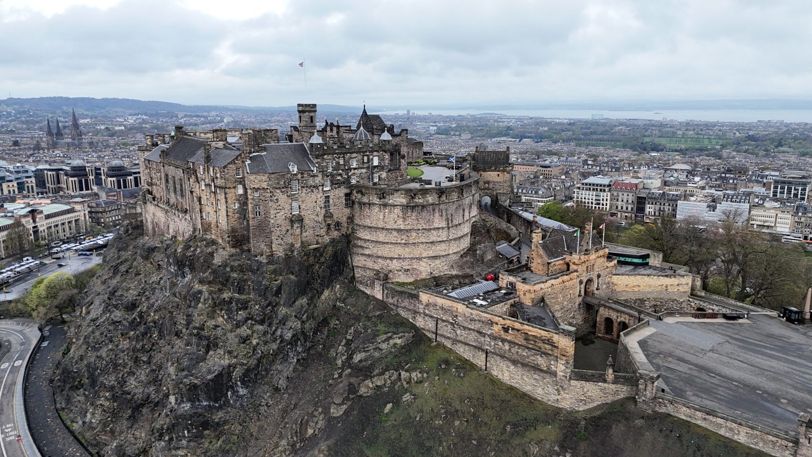 An elevated view of Edinburgh Castle, a large, historic stone fortress with multiple buildings, towers, and walls, perched on a rugged volcanic rock. The castle dominates the left side of the frame. Below and around it, the city of Edinburgh stretches out, with numerous grey buildings and streets. A body of water, likely the Firth of Forth, is visible in the distance. The sky is overcast and grey.
