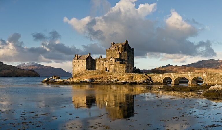 A scenic photograph of Eilean Donan Castle reflected in calm water under a partly cloudy sky at sunset. The historic stone castle sits on a small island connected to the mainland by a stone arch bridge. Mountains with snow-capped peaks are visible in the distance. The water is still, reflecting the warm glow of the sky.