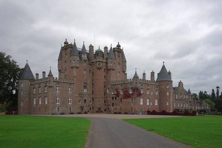 A photo of Glamis Castle, a large, historic Scottish castle with reddish-brown stone walls, numerous turrets, spires, and a central clock tower, under an overcast sky. A paved driveway leads towards the main entrance, flanked by wide green lawns. Some trees with hints of autumn foliage are visible around the castle.