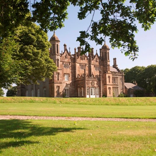 Glenarm Castle: A photo of Glenarm Castle, a large, ornate stone castle with multiple turrets, spires, and intricate architectural details. The castle is framed by lush green trees and sits behind a wide, manicured lawn under a clear blue sky.