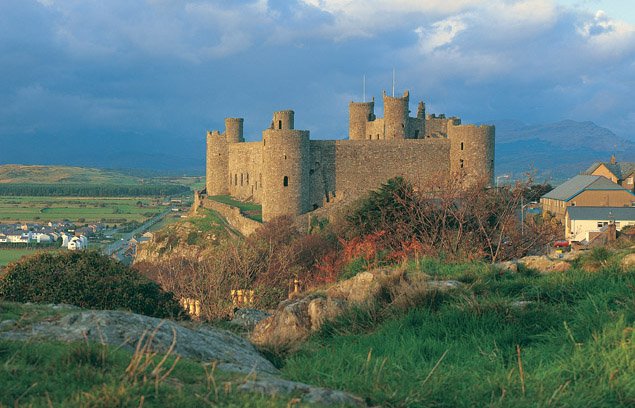An elevated view of Harlech Castle, a large grey stone medieval fortress with multiple round towers and high curtain walls, situated on a dramatic rocky outcrop. The castle dominates the mid-ground, while a town and fields stretch into the distance on the left. The sky above is a dramatic mix of blue and dark clouds, with hints of warm light. The foreground features grassy hills and rocky terrain.