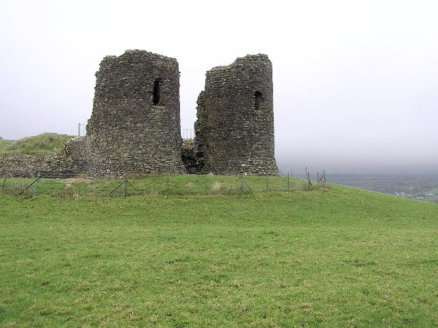 Harry Avery's Castle: A photo of the ruins of Harry Avery's Castle, showing two tall, cylindrical stone towers with jagged tops. The towers are made of dark, rough-cut stone and stand on a grassy hill. The space between the two towers is collapsed, creating a large gap. A wire fence is visible in the foreground, and the background is a misty, grey landscape with a valley below.