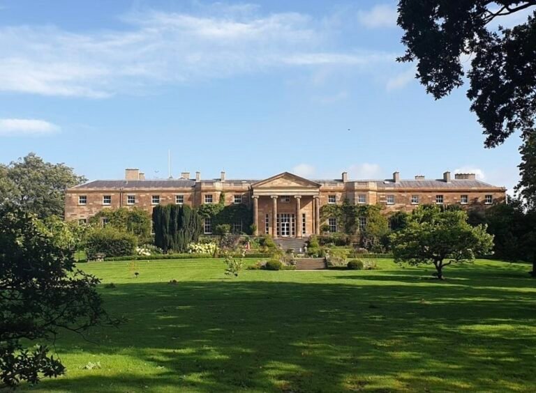 A photo of Hillsborough Castle, a large, two-story Georgian mansion with a symmetrical facade. The building has a central section with a portico and columns, and is constructed from a light-colored stone. The castle is surrounded by manicured green lawns and well-kept gardens with trees and shrubs. The sky is clear and blue with some light clouds.
