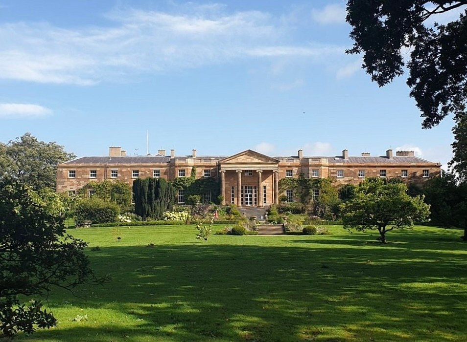 A photo of Hillsborough Castle, a large, two-story Georgian mansion with a symmetrical facade. The building has a central section with a portico and columns, and is constructed from a light-colored stone. The castle is surrounded by manicured green lawns and well-kept gardens with trees and shrubs. The sky is clear and blue with some light clouds.