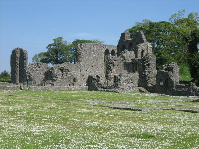 A photo of the extensive stone ruins of Inch Abbey on a sunny day. The fragmented walls show multiple arched window openings and parts of collapsed structures, revealing the sky through them. The foreground is a wide, grassy field dotted with white daisies, and a line of green trees stands behind the ruins.