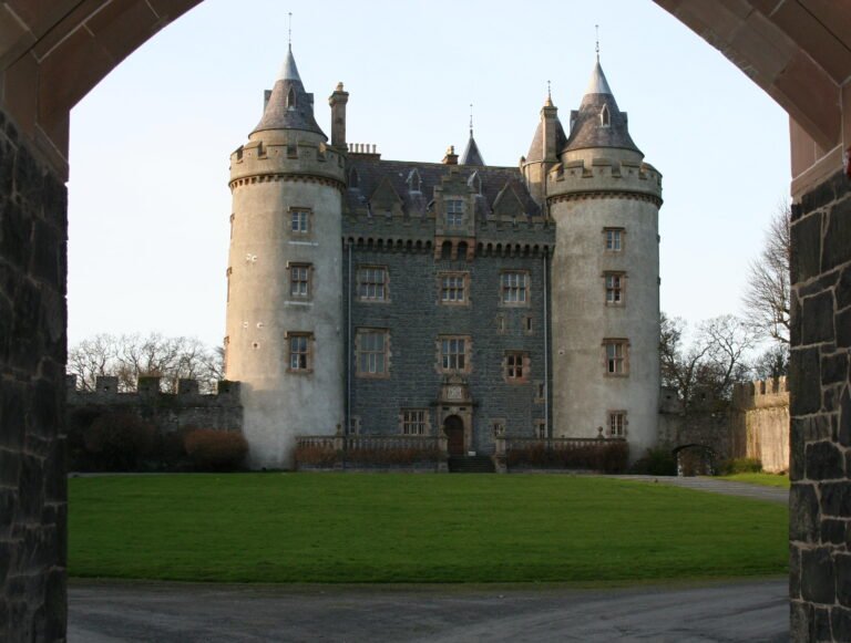 A photo of Killyleagh Castle, a grand, grey stone castle with multiple conical-roofed turrets, viewed through a stone archway. The castle has a central block with numerous windows and a main entrance. A well-maintained green lawn lies in front of the castle, and parts of the exterior wall are visible to the sides. The sky is bright and clear.