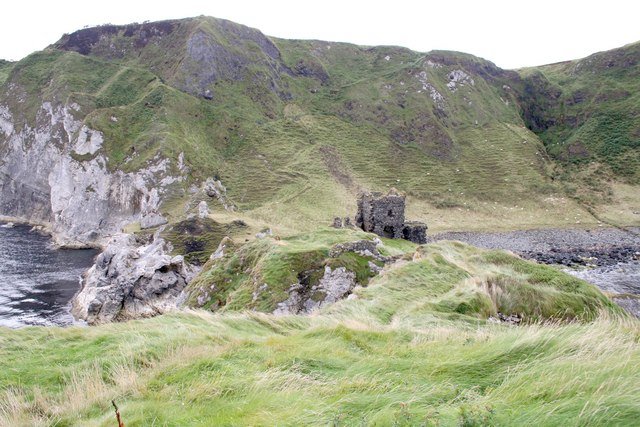 Kinbane Castle: A photo of the ruins of Kinbane Castle, a small stone structure on a narrow, rocky promontory that juts out into the sea. The ruin is surrounded by green grass and rocky outcrops. The promontory is at the base of a large, steep, green cliff. The sea is visible to the left and right.