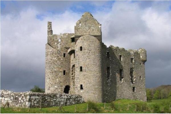 Monea Castle: A photo of the stone ruins of Monea Castle on a sunny day with a dramatic sky. The castle is a large, rectangular structure with two prominent round towers on one side and a taller, gabled section in the center. The stone walls are mostly intact, but the interior is open to the sky, and many windows are empty. A low stone wall extends from the base of the castle in the foreground, and there is green grass around the ruins. The sky is partly cloudy, with bright patches and darker, stormy-looking clouds.