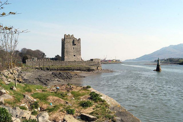 Narrow Water Castle: A photo of Narrow Water Castle, a four-story, grey stone tower with battlements. It stands on a rocky, low-tide shore next to a wide river or inlet. A low stone wall extends from the base of the tower. A small, statue-like marker is visible in the middle of the water. The surrounding landscape is green and hilly, and the sky is clear and bright.