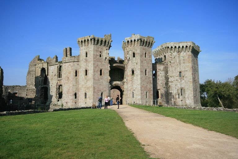 A photo of the impressive main entrance of Raglan Castle, a large stone ruin with two prominent, well-preserved towers flanking a central arched gateway. The towers feature battlements and multiple small window openings. A dirt path leads towards the entrance, surrounded by green grass. A few people are walking near the gateway. The sky is clear and blue.