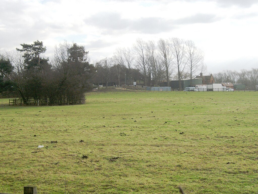 Risinghoe Castle: A wide, grassy field with a line of trees and a building in the background, where the low earthwork mound of Risinghoe Castle is visible.