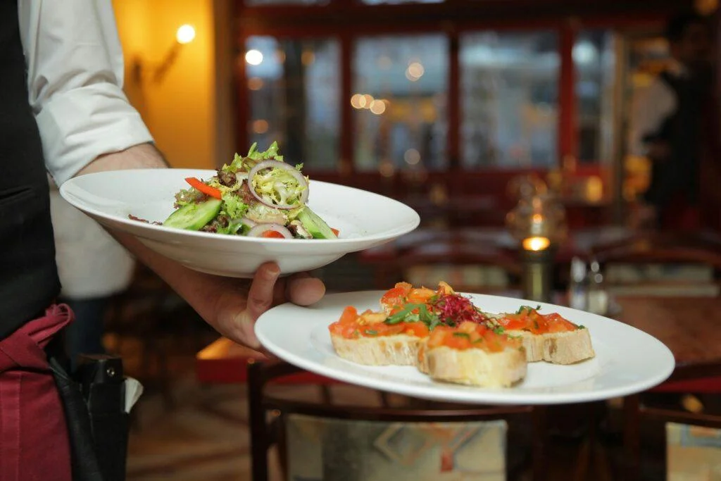 A person in a black vest and white shirt holds two plates of food: a green salad on one plate and an appetizer of bruschetta on the other.
