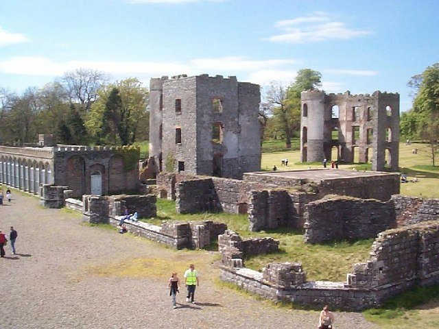 Shane's Castle: A photo of the ruins of Shane's Castle, with several large, fragmented stone towers and walls scattered across a grassy field. The main ruined structures are two large, grey stone buildings with gaping window openings and missing roofs. A long, arched colonnade extends to the left. A few people are seen walking on the dirt ground and sitting on low walls among the ruins. The sky is bright and blue with white clouds.