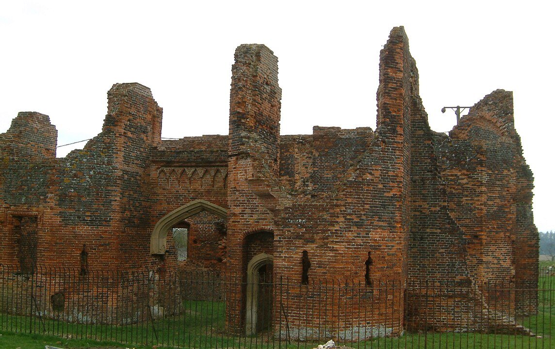Someries Castle: The brick ruins of Someries Castle Chapel, showing a section of weathered walls and a large arched window with a metal fence in the foreground.
