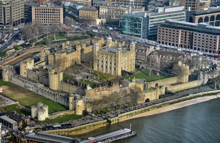 An aerial view of the Tower of London, showing its central White Tower and surrounding walls, with the River Thames and a modern cityscape in the background.