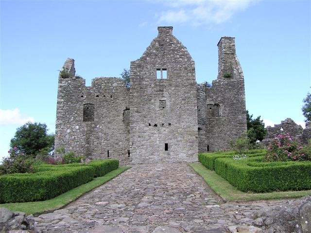 Tully Castle: A photo of the ruins of Tully Castle, a grey stone structure with a tall, gabled central section and fragmented side walls, viewed from a cobbled pathway. The pathway is flanked by neatly trimmed green hedges and flower beds. The castle walls have empty window openings, and parts of the upper structure are missing. The sky is bright blue with a few white clouds.