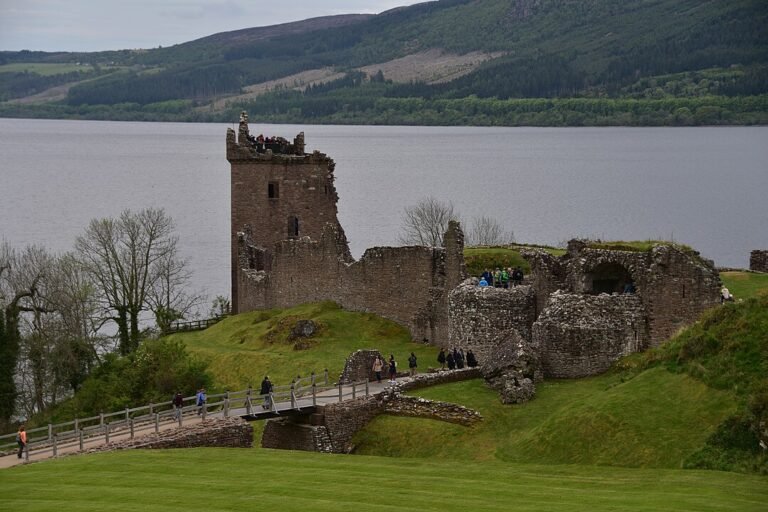 A scenic view of the stone ruins of Urquhart Castle on the shore of a wide, calm loch under an overcast sky. A prominent, tall rectangular tower with battlements and missing sections stands to the left. To its right, various fragmented stone walls and mounds are scattered across grassy slopes. A wooden walkway with handrails leads towards the ruins, with several people walking on it and exploring the site. In the background, the opposite shore of the loch is lined with dark green forested hills.