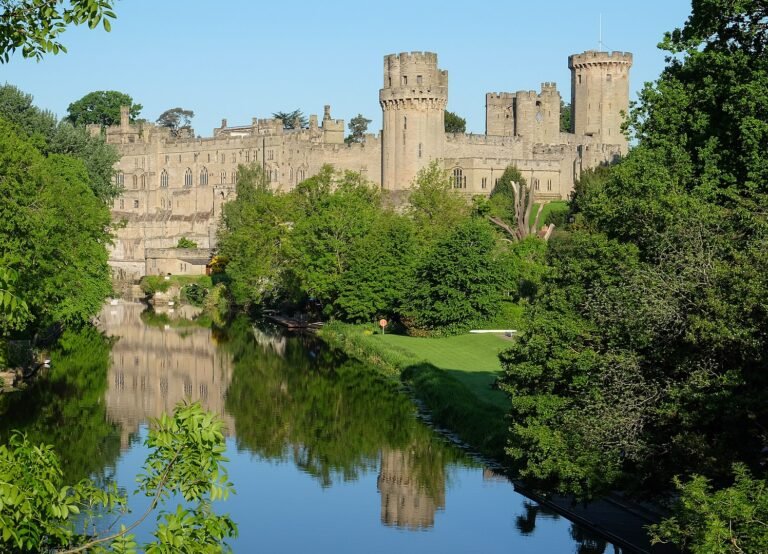 Warwick Castle's towers and stone walls reflected in the calm waters of the River Avon, surrounded by lush green trees under a clear blue sky.