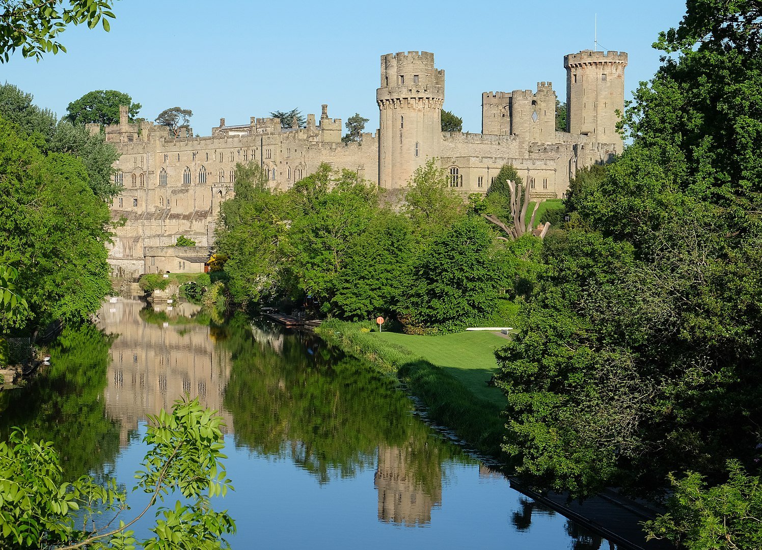 Warwick Castle's towers and stone walls reflected in the calm waters of the River Avon, surrounded by lush green trees under a clear blue sky.