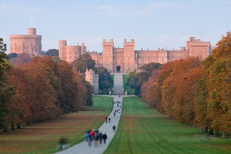 An eye-level shot of Windsor Castle's main buildings and a large round tower, seen from a long, tree-lined path with people walking towards the castle during sunset.