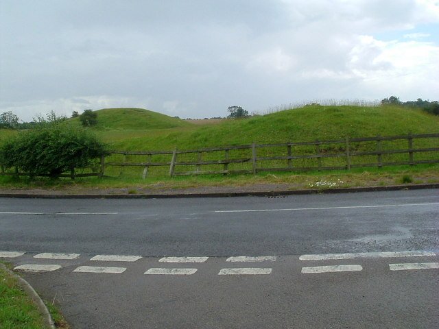 Yielden Castle: The remains of a motte-and-bailey castle, with two large, grassy mounds separated by a fence from a paved road.