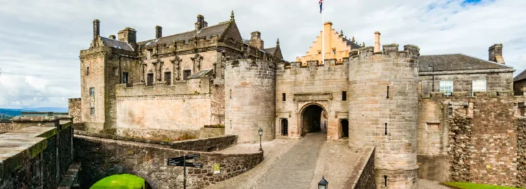 A panoramic view of the entrance to Stirling Castle, a grand stone fortress with multiple buildings, towers, and a prominent gatehouse. A long, paved pathway leads up to an arched entrance flanked by round towers. To the left, a large, ornate building with many windows and decorative elements is visible. The sky is cloudy, and a Scottish flag flies above one of the castle sections.